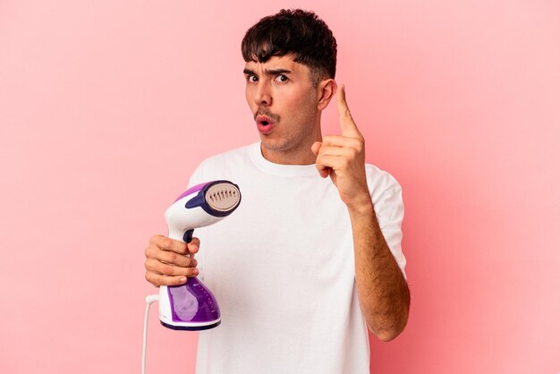 Homem jovem de raça mista segurando um ferro isolado no fundo rosa, tendo uma ideia, conceito de inspiração.