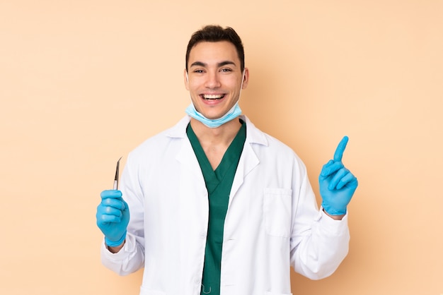 Homem jovem dentista segurando ferramentas na parede bege apontando uma ótima idéia