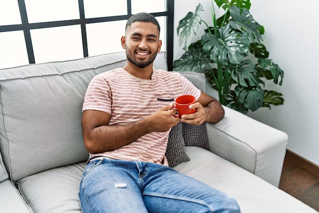 Homem jovem do Oriente Médio sorrindo confiante bebendo café em casa