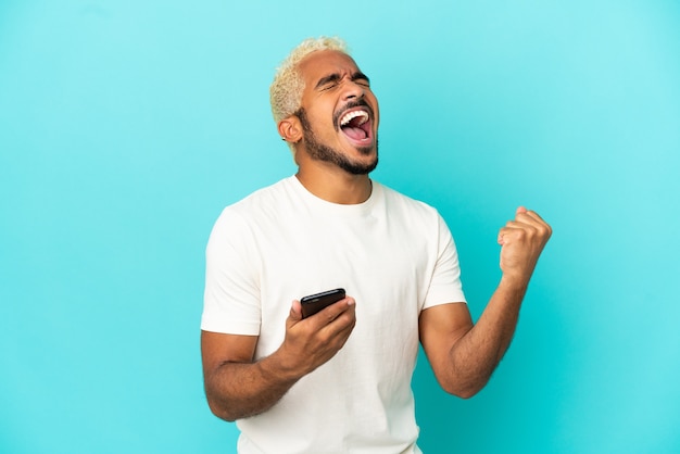 Homem jovem e bonito colombiano isolado em um fundo azul com o telefone em posição de vitória