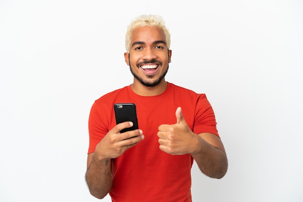 Homem jovem e bonito colombiano isolado no fundo branco, usando um telefone celular enquanto faz o polegar para cima