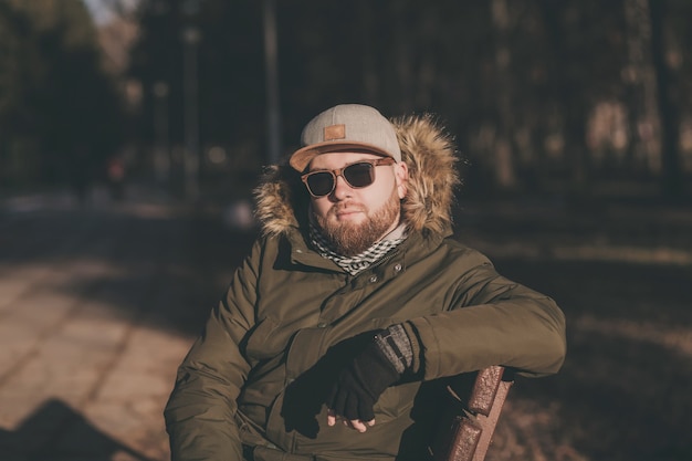 Homem jovem e bonito hippie em óculos de sol sentado em um banco ao ar livre