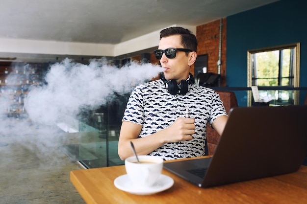 Homem jovem e bonito hipster em óculos de sol sentado no café com uma xícara de café vaping e libera um cl