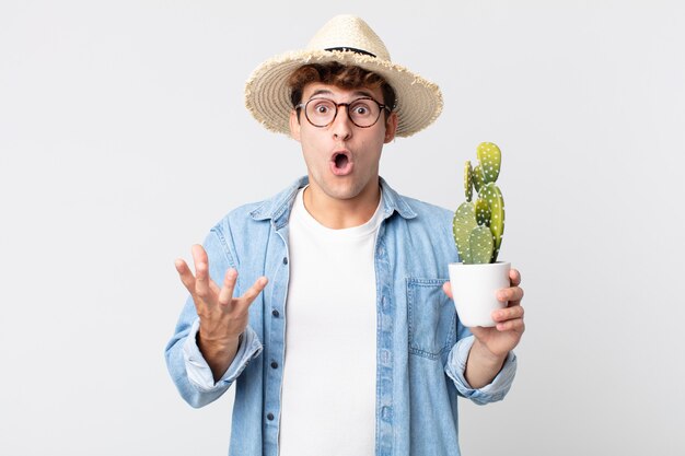 Homem jovem e bonito maravilhado, chocado e atônito com uma surpresa inacreditável. fazendeiro segurando um cacto decorativo