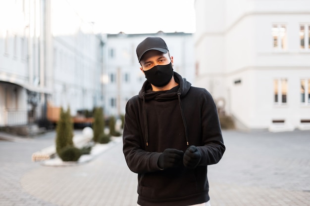 Homem jovem e bonito modelo com máscara médica protetora e boné elegante com capuz preto na moda e luvas na cidade. Estilo urbano moderno masculino e pandemia