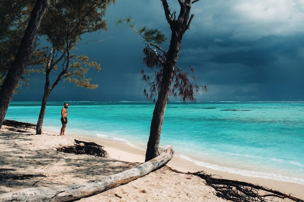 Homem jovem e bonito relaxado em shorts preto, em pé na praia.