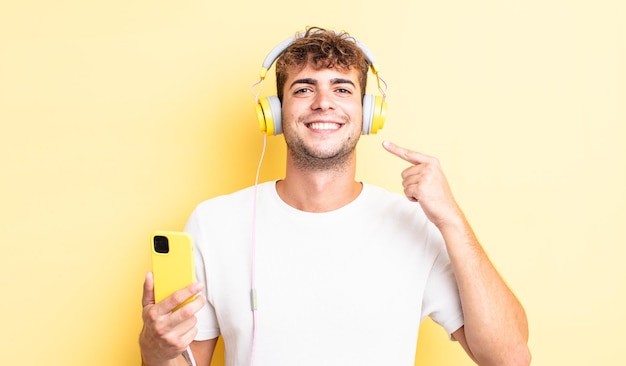 Homem jovem e bonito sorrindo com confiança, apontando para o próprio sorriso largo. conceito de fones de ouvido e smartphone