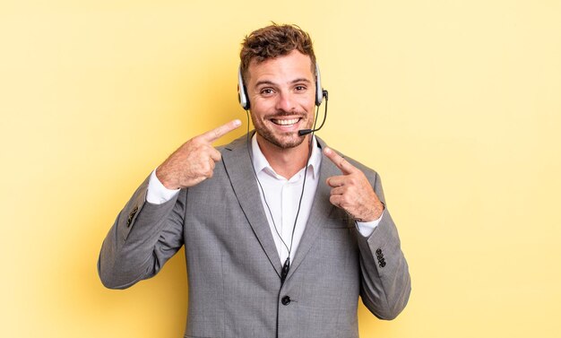 Homem jovem e bonito sorrindo com confiança, apontando para o próprio sorriso largo. concetp de telemarketing