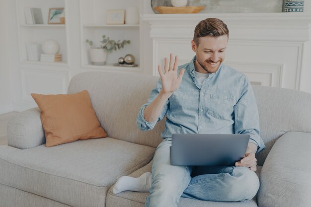 Homem jovem e bonito vestido de maneira casual acenando com a mão enquanto fala com a família online usando um notebook, feliz por ter uma conversa com parentes, sentado no sofá com as pernas cruzadas