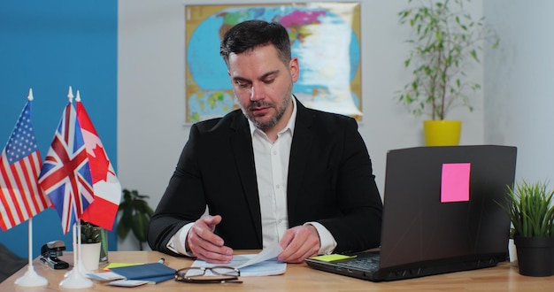 Homem jovem e elegante trabalha em uma agência de viagens no computador portátil sentado em uma mesa no fundo de um mapa do mundo Conceito de viagem