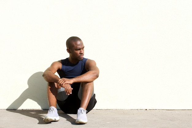 Homem jovem e saudável de esportes afro-americanos sentado ao lado da parede