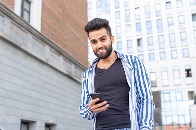 Homem jovem elegante hippie árabe com barba conversando nas redes sociais usando um smartphone e a internet sem fio em um dia quente de verão.