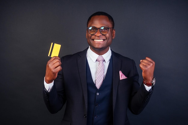 Homem jovem empresário de sucesso em um elegante terno preto clássico e óculos legais, segurando um cartão de crédito de plástico amarelo no estúdio em um fundo escuro. conceito de compras venda de sexta feira negra