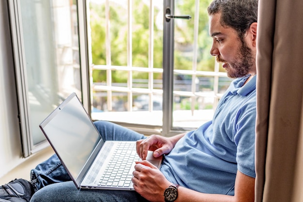 Homem jovem empresário usando computador portátil sentado ao lado da janela do apartamento