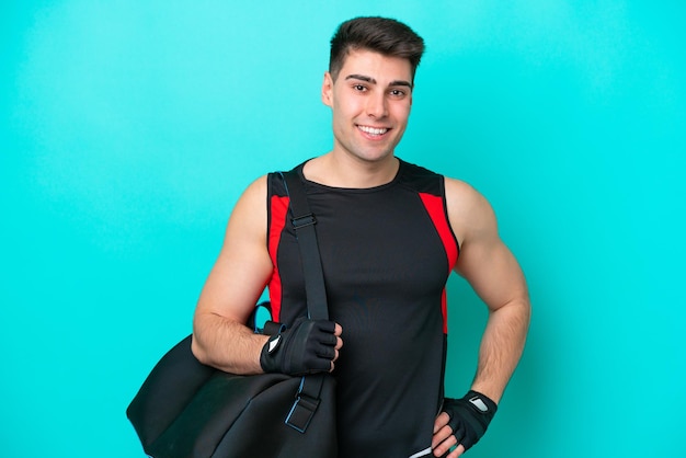 Homem jovem esporte caucasiano com bolsa esportiva isolada em fundo azul posando com braços no quadril e sorrindo