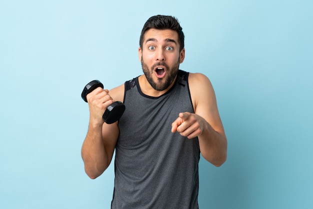 Homem jovem esporte com barba fazendo levantamento de peso surpreso e apontando a frente