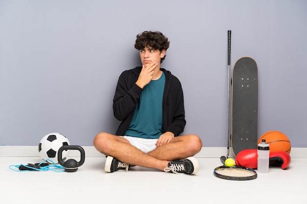 Homem jovem esporte sentado no chão em torno de muitos elementos de esporte, pensando uma idéia