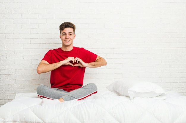 Homem jovem estudante adolescente na cama sorrindo e mostrando uma forma de coração com as mãos.