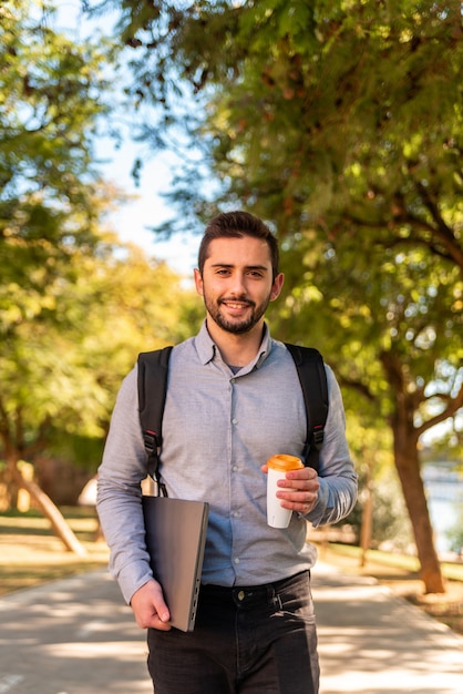 Homem jovem estudante e trabalhador caucasiano, bebendo um café para viagem e carregando seu laptop em um belo parque em um dia ensolarado.