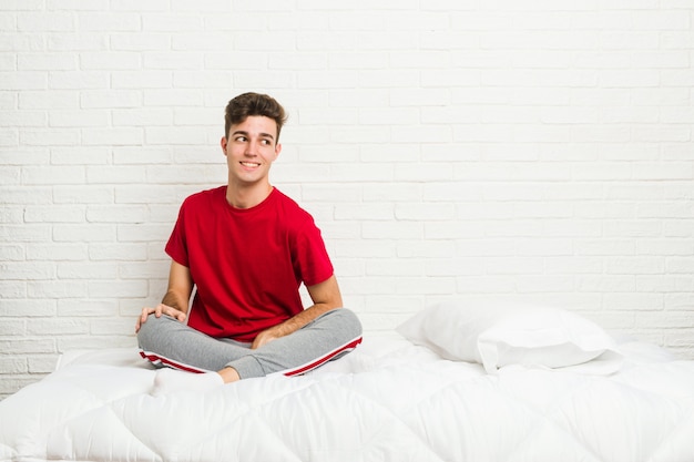 Homem jovem estudante estudante na cama parece de lado sorrindo, alegre e agradável.