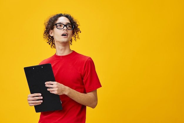 Homem jovem estudante míope confuso em óculos engraçados de camiseta vermelha detém pasta de tablet com notas de estudo parece de lado posando isolado sobre fundo amarelo Lugar livre para anúncio Conceito de faculdade de educação