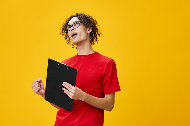 Homem jovem estudante míope feliz alegre em óculos engraçados de camiseta vermelha detém pasta de tablet com notas de estudo posando isoladas sobre fundo amarelo do estúdio Lugar livre para anúncio Conceito de faculdade de educação