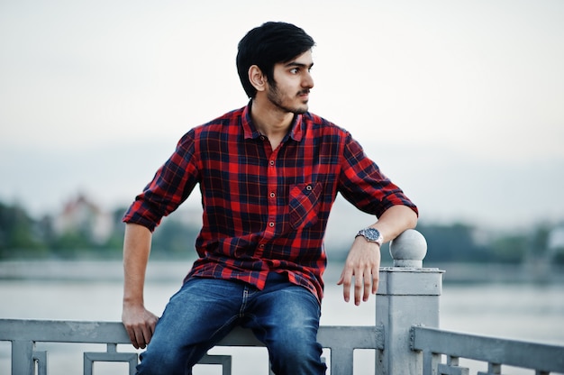 Homem jovem estudante na camisa quadriculada e calça jeans, sentado nos corrimãos contra o lago