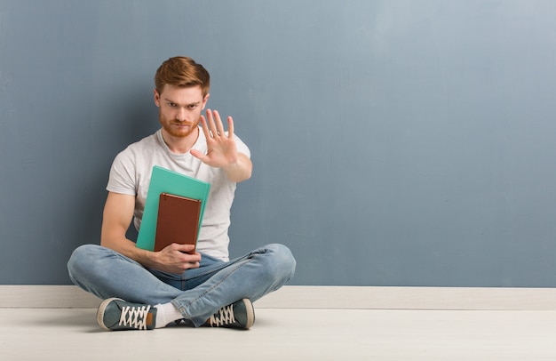 Homem jovem estudante ruiva sentada no chão, colocando a mão na frente. ele está segurando livros.