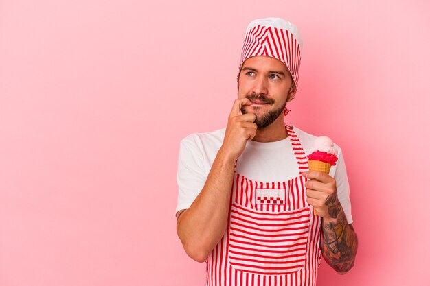 Homem jovem fabricante de gelo caucasiano com tatuagens segurando sorvete isolado no fundo rosa relaxado pensando em algo olhando para um espaço de cópia.