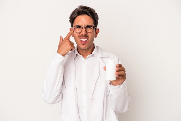 Homem jovem farmacêutico raça mista segurando comprimidos isolados no fundo branco, mostrando um gesto de decepção com o dedo indicador.