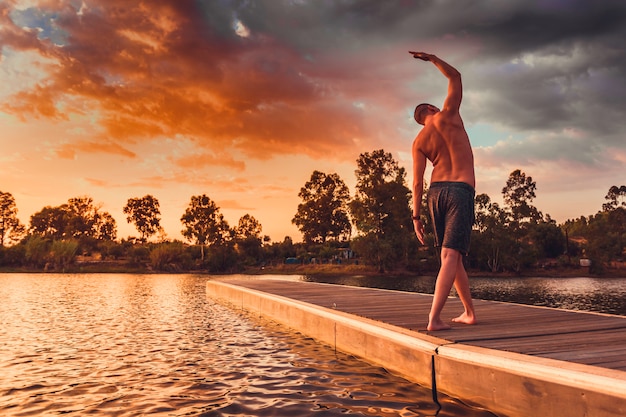 Homem jovem, fazendo, pilates, exercícios, ficar, ligado, madeira, jetty, em, pôr do sol