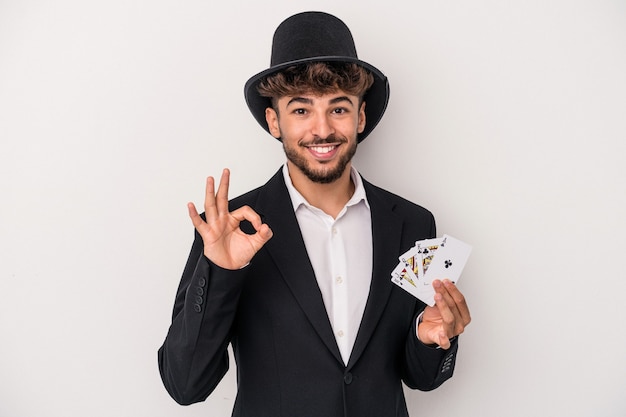 Homem jovem feiticeiro árabe segurando cartas mágicas isoladas no fundo branco, alegre e confiante, mostrando um gesto ok.