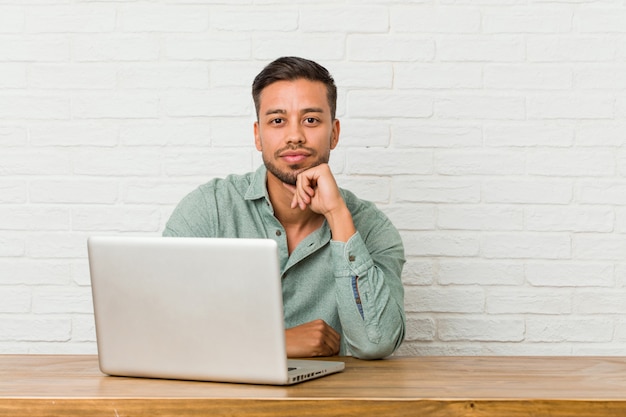 Homem jovem filipino sentado trabalhando com seu laptop sorrindo feliz e confiante, tocando o queixo com a mão