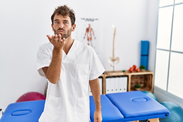 Homem jovem fisioterapeuta bonito trabalhando na clínica de recuperação de dor olhando para a câmera mandando um beijo com a mão no ar sendo adorável e sexy expressão de amor