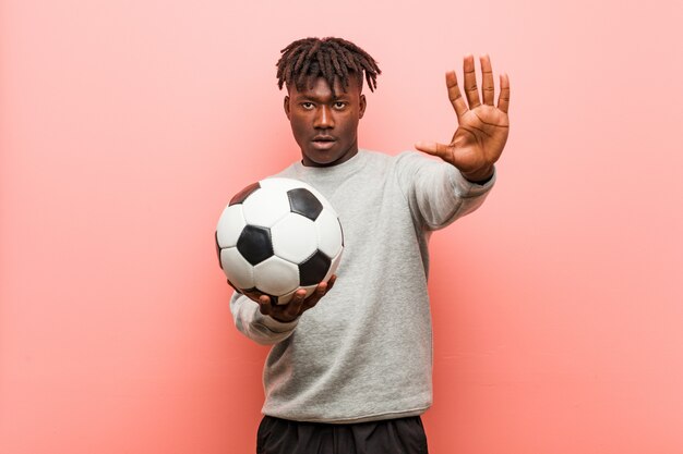 Homem jovem fitness preto segurando uma bola de futebol em pé com a mão estendida, mostrando o sinal de stop, impedindo-o.