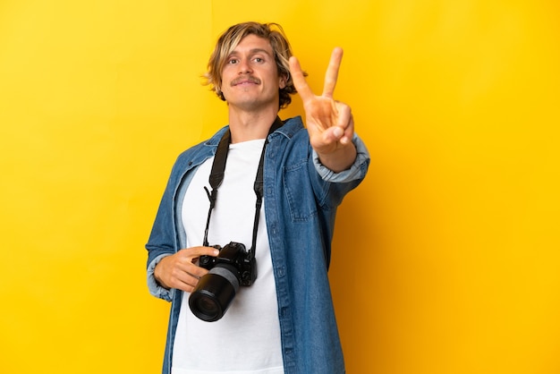 Homem jovem fotógrafo isolado na parede amarela sorrindo e mostrando sinal de vitória