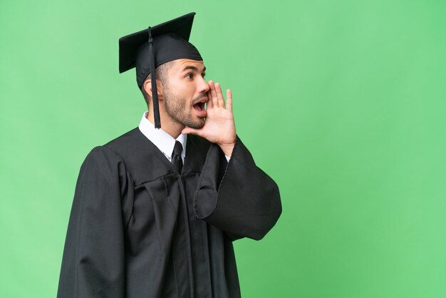 Homem jovem graduado da universidade sobre fundo isolado, gritando com a boca aberta para o lado