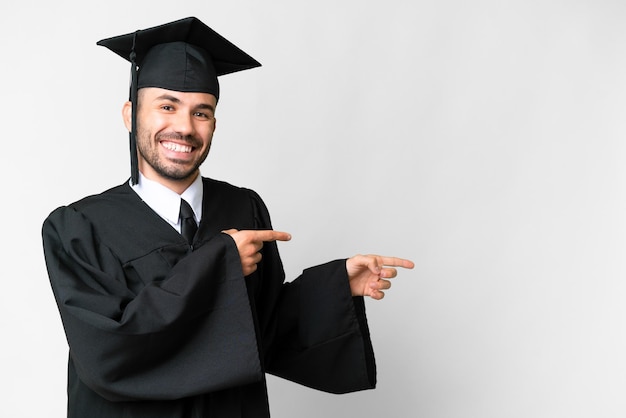 Homem jovem graduado universitário sobre fundo branco isolado, apontando o dedo para o lado e apresentando um produto