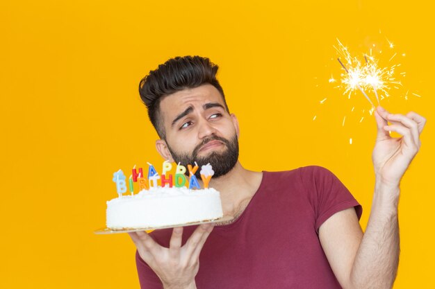 Homem jovem hippie envergonhado segura duas velas acesas e um bolo de felicitações, posando sobre um fundo amarelo. conceito de férias sem fim.