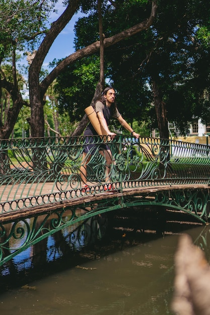 Homem jovem hippie latino com cabelos longos, apoiando-se no corrimão de uma ponte de pedestres, olhando para o horizonte Foto vertical