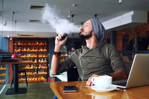 Homem jovem hipster bonito com barba sentado no café com uma xícara de café vaping e libera um clo