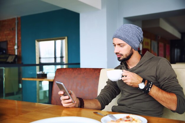 Homem jovem hipster bonito com barba sentado no café usando um telefone celular segurando uma xícara de café