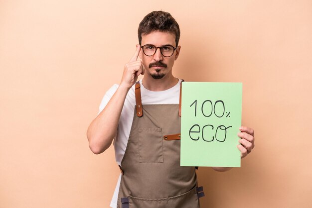 Homem jovem jardineiro caucasiano segurando 100% eco cartaz isolado em fundo bege, apontando o templo com o dedo, pensando, focado em uma tarefa.