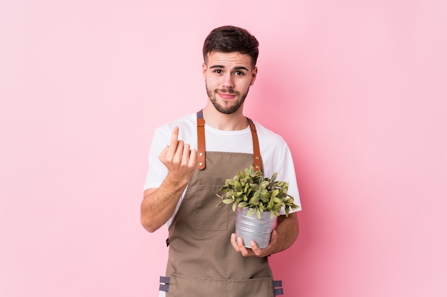 Homem jovem jardineiro caucasiano segurando uma planta apontando com o dedo para você, como se convidando se aproximar.