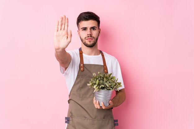 Homem jovem jardineiro caucasiano segurando uma planta isolada em pé com a mão estendida, mostrando o sinal de stop, impedindo-o.
