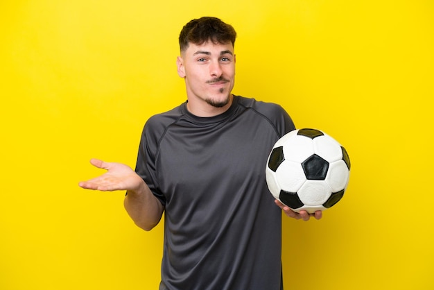 Homem jovem jogador de futebol isolado em fundo amarelo com dúvidas ao levantar as mãos