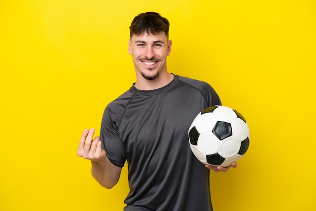 Homem jovem jogador de futebol isolado em fundo amarelo, fazendo gesto de dinheiro