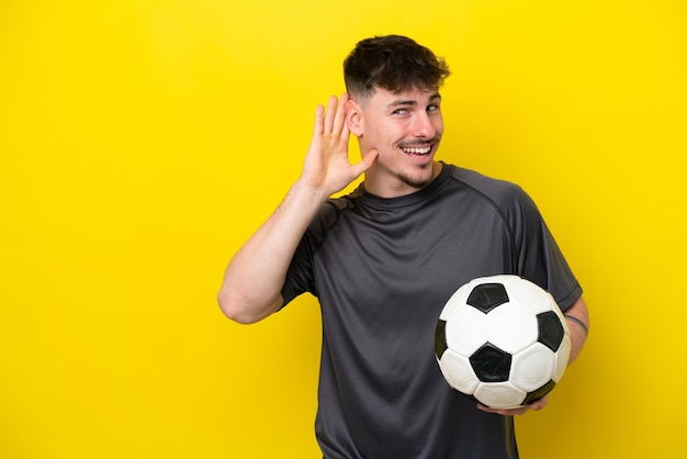 Homem jovem jogador de futebol isolado em fundo amarelo ouvindo algo colocando a mão na orelha