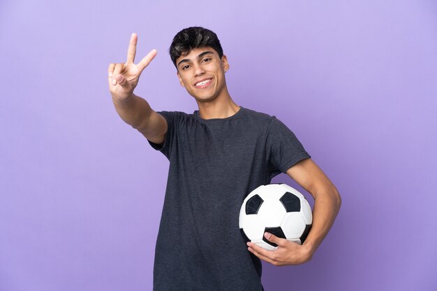 Homem jovem jogador de futebol sobre um roxo isolado sorrindo e mostrando sinal de vitória