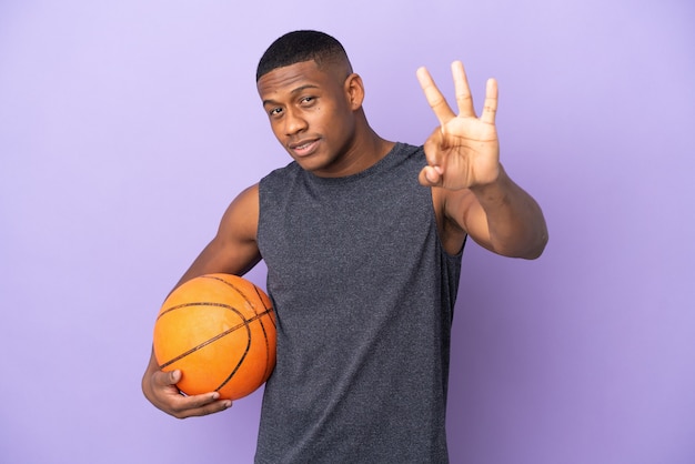 Homem jovem jogador latino de basquete isolado na parede roxa feliz e contando três com os dedos